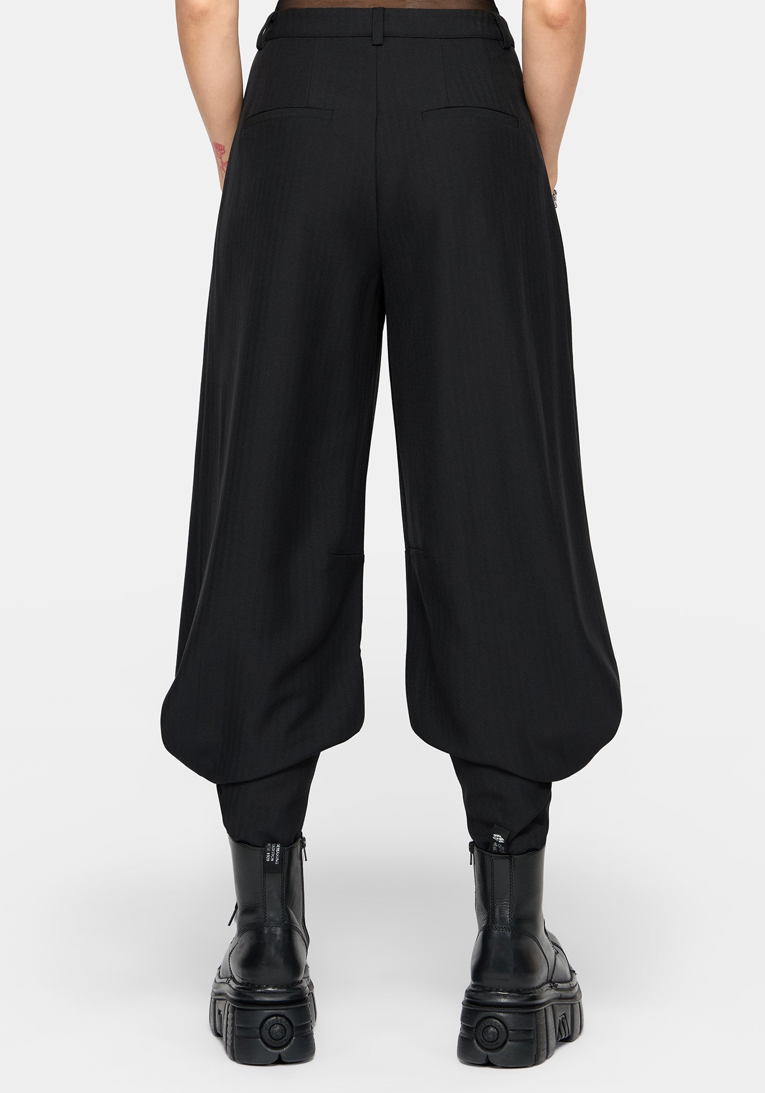 Rear view of a model wearing black wide-leg balloon pants with gathered legs and black platform ankle boots against a light gray studio background.