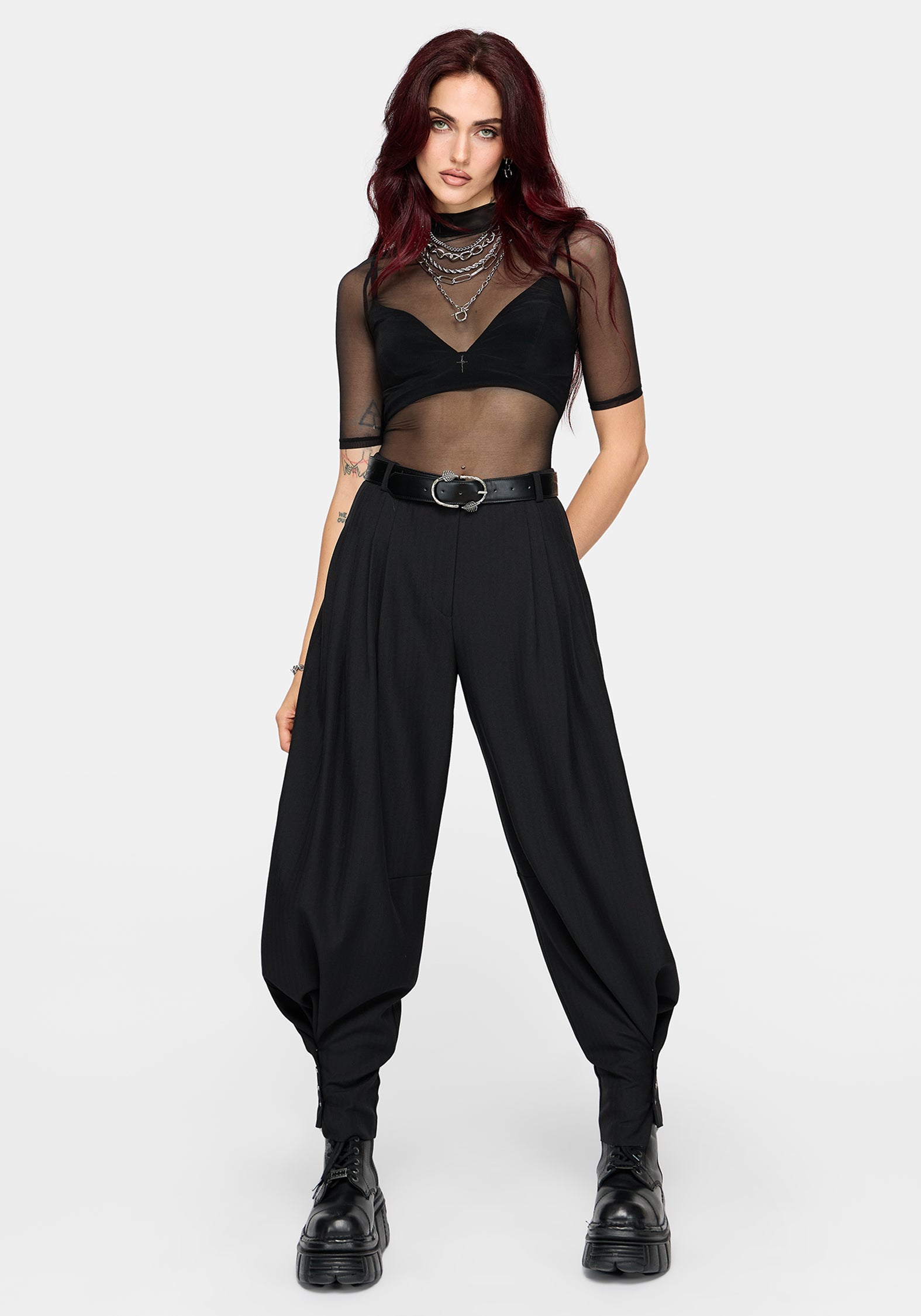 A model with dark red hair wearing a black sheer mesh short-sleeve crop top, black high-waisted balloon pants, a black belt, and black boots, standing against a light gray studio background.