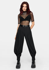 A model with dark red hair wearing a black sheer mesh short-sleeve crop top, black high-waisted balloon pants, a black belt, and black boots, standing against a light gray studio background.